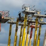 Multiple aerial work platforms with colorful booms reaching skyward against a cloud-dotted backdrop.