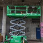Green scissor lift parked outside an urban building with safety cones around.