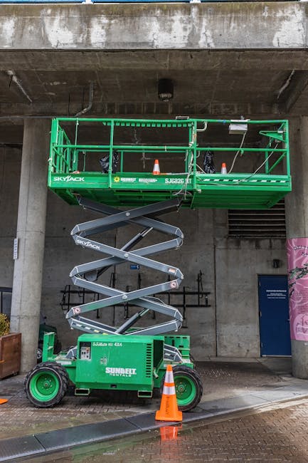 Green scissor lift parked outside an urban building with safety cones around.